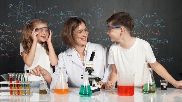 Caucasian boy mixing chemical liquid while teacher giving advice. Professional instructor wearing lab suit looking for diverse student at table with beaker filled with colored solution. Erudition. - Photo, Image