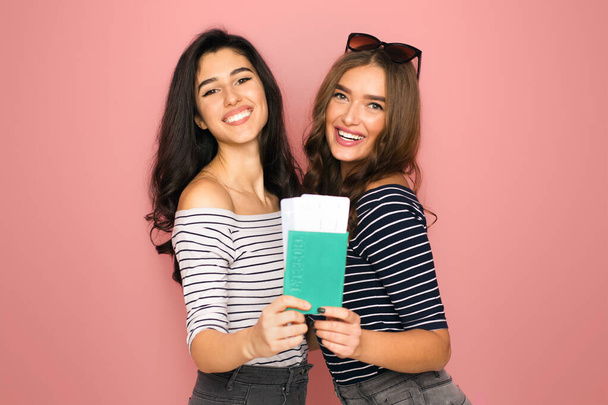 Δύο χαρούμενες νεαρές γυναίκες, φορώντας casual ριγέ μπλούζες, στέκονται η μία κοντά στην άλλη σε ένα ζωντανό ροζ φόντο. Μοιράζονται ένα χαμόγελο ενώ κατέχουν διαβατήριο με εισιτήρια μαζί - Φωτογραφία, εικόνα