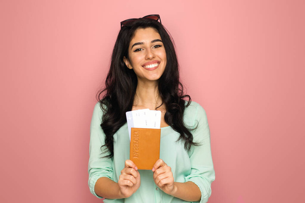 A cheerful young middle eastern woman stands against a vibrant pink backdrop, clutching a passport and a boarding pass, suggesting she is prepared and excited for an upcoming journey - Photo, Image
