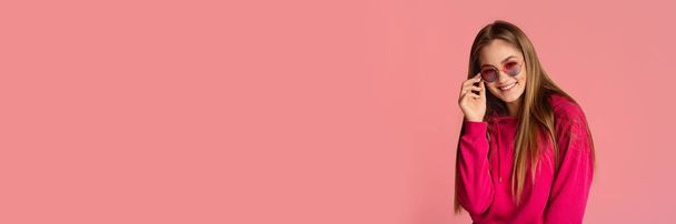 Chica de pelo largo bastante ajustando gafas de sol con estilo, posando aislado sobre fondo de estudio rosa, sonriendo a la cámara, panorama con espacio de copia - Foto, imagen