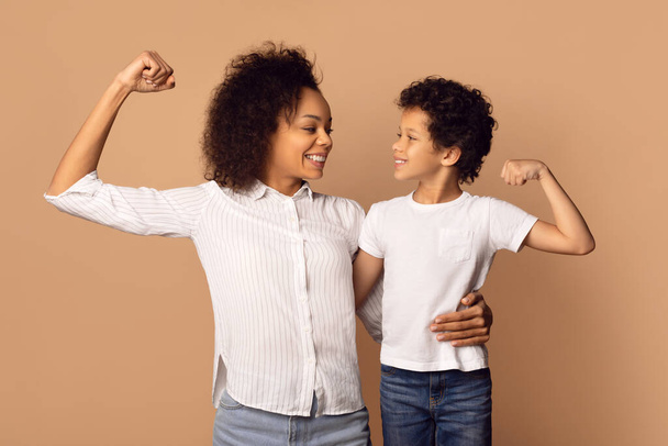 Afro-Amerikaanse vrouw en een jongen staan naast elkaar, tonen biceps en glimlachen, geïsoleerd op beige achtergrond - Foto, afbeelding
