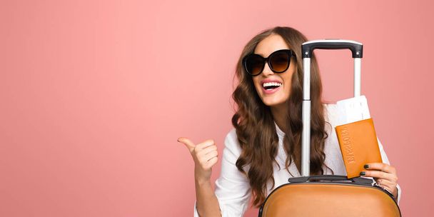 Μια χαρούμενη νεαρή γυναίκα φορώντας μεγάλα γυαλιά ηλίου στέκεται με τις αποσκευές της, δίνοντας μια πινακίδα αντίχειρες προς τα πάνω. Είναι ντυμένη άνετα, έτοιμη για ένα άνετο και κομψό ταξίδι, αντίγραφο χώρου - Φωτογραφία, εικόνα
