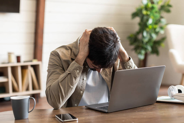 Egy fiatalember ül egy fa íróasztalnál, a kezei szorongatják a fejét egy gesztussal a frusztráció vagy túlterheltség. Nyílt laptop, mobiltelefon és egy csésze kávé kíséri. - Fotó, kép