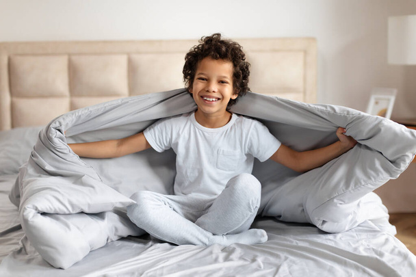 Der afroamerikanische Junge mit dem lockigen Haar sitzt auf einem ordentlich gemachten Bett, sein ansteckendes Lächeln strahlt Freude aus, als er in einem sonnendurchfluteten Schlafzimmer spielerisch eine weiche graue Decke hochhält. - Foto, Bild