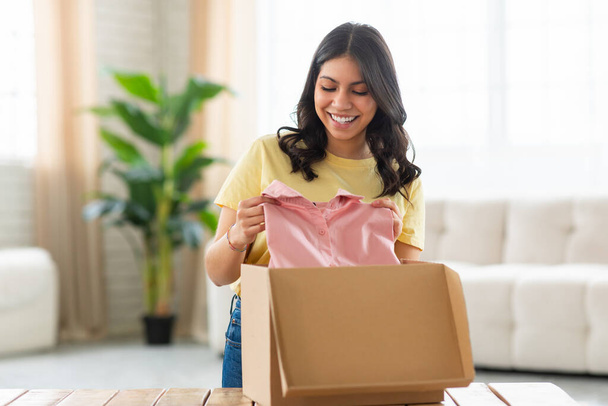 A delighted young middle eastern woman is standing in a well-lit living room, gently pulling out a pink article of clothing from an open cardboard packaging box that rests on a wooden table - Photo, Image