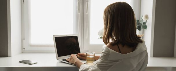 Lähikuva takaisin näkymä nuori melko keskittynyt nainen kirjoittamalla ja selaamalla verkossa kannettavan tietokoneella työskennellessään viihtyisässä huoneessa yksin. Etäopiskelun ja työn käsite. - Valokuva, kuva