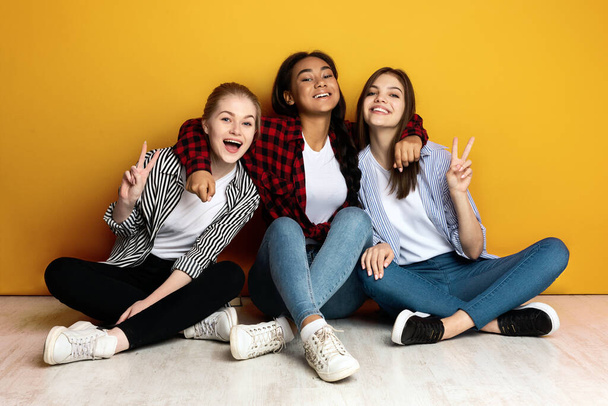 Tre giovani ragazze multietniche siedono fianco a fianco con le spalle contro un muro giallo brillante. Sono vestiti con disinvoltura in jeans e camicie di flanella e sorridono gioiosamente. - Foto, immagini