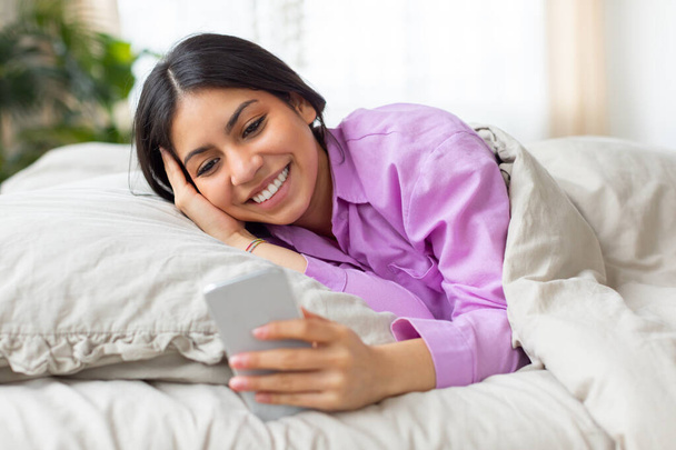Een vrolijke jonge vrouw uit het Midden-Oosten ligt comfortabel in bed, met een roze shirt aan, terwijl ze geniet van het surfen op haar smartphone in de zacht verlichte rust van de ochtend. - Foto, afbeelding