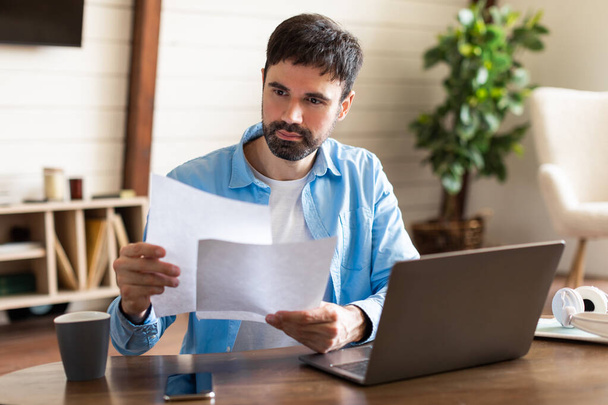 Un hombre está parado y sosteniendo un pedazo de papel delante de un portátil. El hombre aparece enfocado mientras mira la pantalla del portátil y sostiene el papel - Foto, imagen