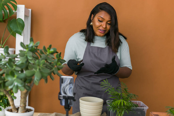 African american girl vlogger influencer or SME owner work on home video camera selfie shoot filming take care home plants and transplanting plant in flowerpot. Home gardening and florist - Foto, imagen