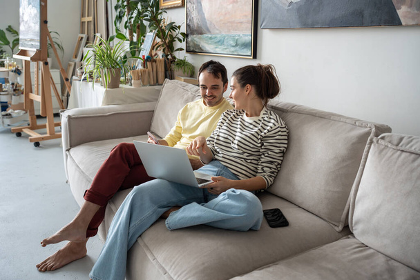 Kreatív nyugodt boldog pár ül kényelmes kanapén laptoppal otthon művészeti stúdió. Mosolygó nő művész megvitatni férfi együttműködési projekt, pihenés kézműves műhelyben. Csapatmunka ötletbörze. - Fotó, kép