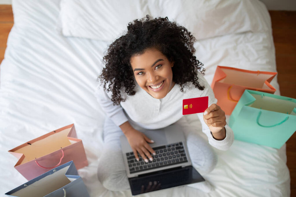 Nainen istuu sängyllä luottokortti toisessa kädessä ja kannettava tietokone toisessa kädessä. Hän näyttää keskittynyt ja harjoittaa online-liiketoimen tai rahoitustoiminnan, ylhäältä - Valokuva, kuva
