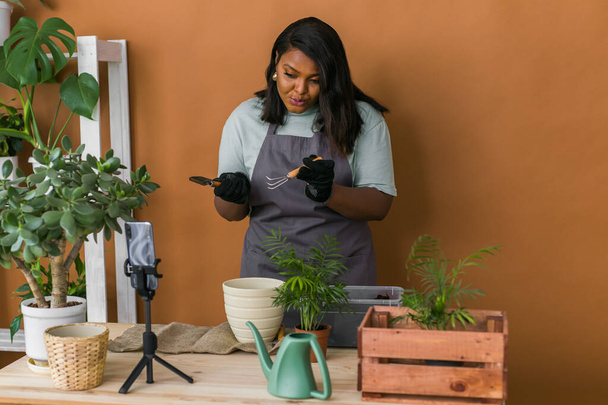 Afrykańska dziewczyna vlogger influencer lub właściciel MŚP pracy na domu aparat wideo selfie sesji filmowej dbać o rośliny domowe i roślin transplantacyjnych w doniczce. Ogrodnictwo i kwiaciarnia - Zdjęcie, obraz