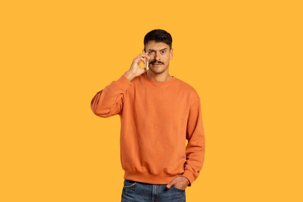 Un hombre que usa un suéter naranja está involucrado en una conversación telefónica usando un teléfono celular sobre fondo amarillo. - Foto, imagen