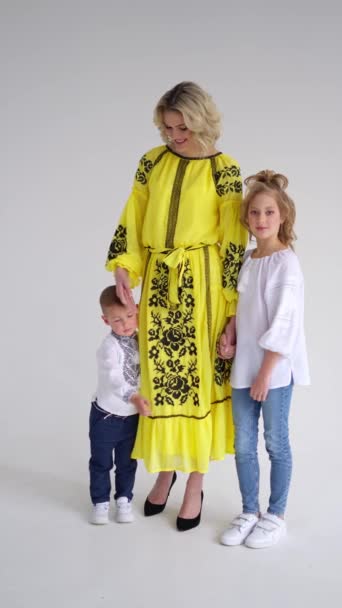 Μητέρα με φωτεινά ρούχα κοιτάζει τα παιδιά της: κορίτσι με λευκό φόρεμα και αγόρι με κεντημένο πουκάμισο στέκονται το ένα δίπλα στο άλλο, αποδεικνύοντας την οικογενειακή αρμονία. - Πλάνα, βίντεο