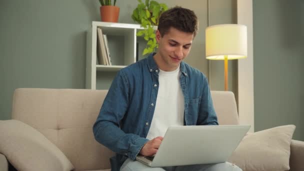 Gelukkige freelancer die overdag thuis werkt met een laptop op een bank. Jongeman glimlachen en surfen op internet met behulp van een computer in de woonkamer. Ondernemer op zoek bank financiert en investeert - Video