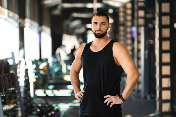 Ένας άντρας στέκεται με αυτοπεποίθηση σε ένα γυμναστήριο, με τα χέρια του να ακουμπάνε στους γοφούς του. Φαίνεται συγκεντρωμένος και αποφασισμένος, έτοιμος για την προπόνηση.. - Φωτογραφία, εικόνα