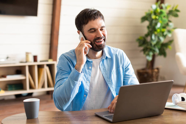 Ein Mann im blauen Hemd sitzt an einem Holztisch und telefoniert mit seinem Handy. Der Mann wirkt konzentriert, als er ins Telefon spricht, sein Gesichtsausdruck ernst.. - Foto, Bild