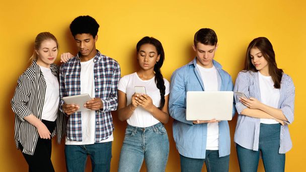 Πέντε πολυεθνικοί έφηβοι στέκονται δίπλα-δίπλα, ο καθένας απορροφημένος σε διαφορετικές ηλεκτρονικές συσκευές, ποζάροντας σε κίτρινο φόντο στούντιο. - Φωτογραφία, εικόνα