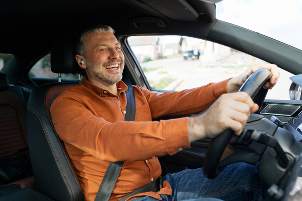 Glimlachende Europese man of bestuurder van middelbare leeftijd met draadloze oortelefoons die auto rijden en genieten van muziek - Foto, afbeelding