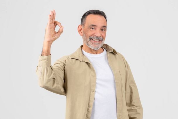 Un uomo anziano che indossa una camicia bianca e giacca abbronzata è visto fare il segno Ok con la mano. Le sue dita sono divise nell'iconica forma OK - Foto, immagini