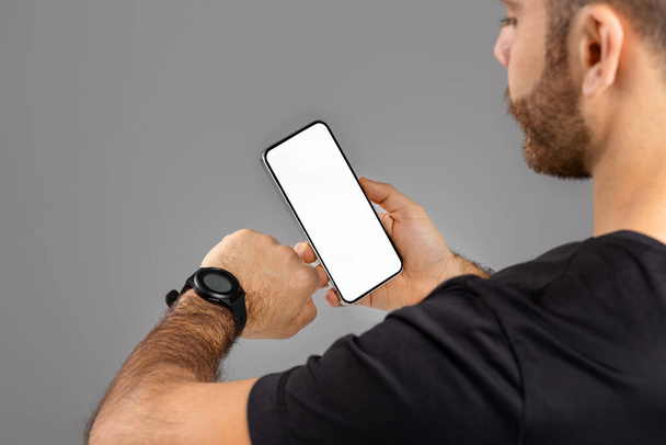 Um homem é visto de lado enquanto levanta o braço esquerdo, olhando para seu smartwatch, enquanto segura um smartphone com uma tela em branco na mão direita, rastreando seu treino - Foto, Imagem