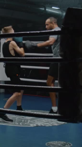 Vertical shot of Caucasian boy in boxing gloves practices in fighting technique, trains with African American trainer on boxing ring. Teen hits punching mitts and exercise in dark gym. - Footage, Video