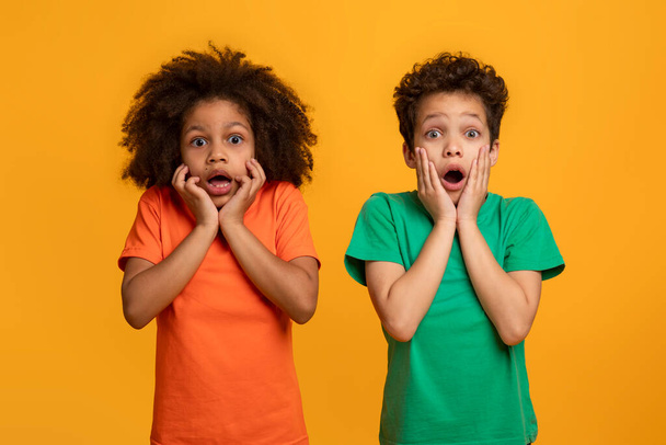 Two young African American children stand side by side, their eyes wide and hands on their cheeks in a classic expression of surprise and astonishment. The vibrant yellow backdrop - Photo, Image