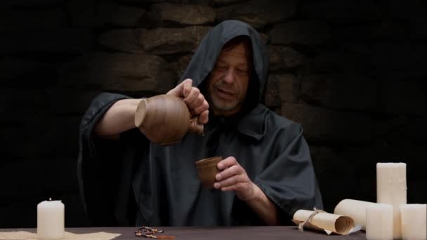 Een monnik in een zwarte soutane schenkt en drinkt wijn terwijl hij aan een tafel tegen een stenen muur zit. - Video