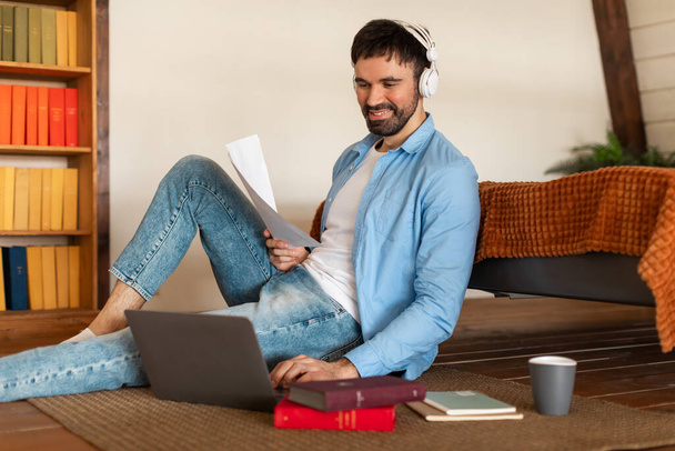 Een jongeman op een houten vloer leunt tegen een bank, glimlachend terwijl hij een document leest en naar muziek luistert via zijn koptelefoon. - Foto, afbeelding