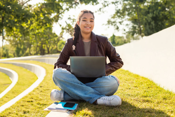 Una estudiante está sentada en el suelo, trabajando en un portátil. Aparece enfocada y comprometida en su trabajo, rodeada de un trasfondo neutral.. - Foto, imagen