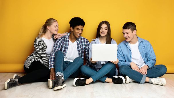Vier multi-etnische tieners zitten terloops op de vloer met hun rug naar een levendige gele muur. Een van hen houdt een open laptop vast en laat iets interessants zien. - Foto, afbeelding