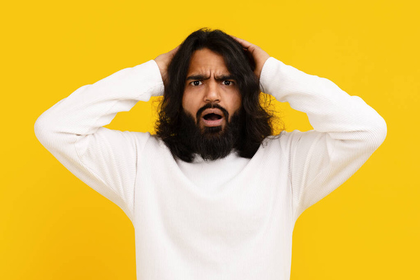 Indiase man met lang haar en een baard staat tegen een levendige gele achtergrond, zijn uitdrukking van intense frustratie, het aanraken van zijn hoofd - Foto, afbeelding