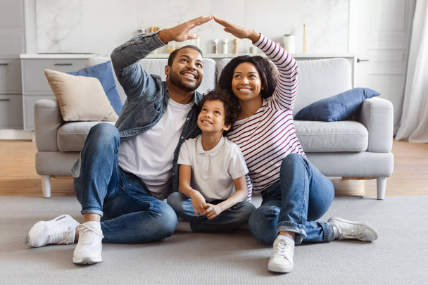 Une joyeuse famille afro-américaine est assise sur le sol du salon avec un canapé en arrière-plan. L'homme et la femme souriants lèvent leurs bras pour former un toit au-dessus de leur enfant - Photo, image