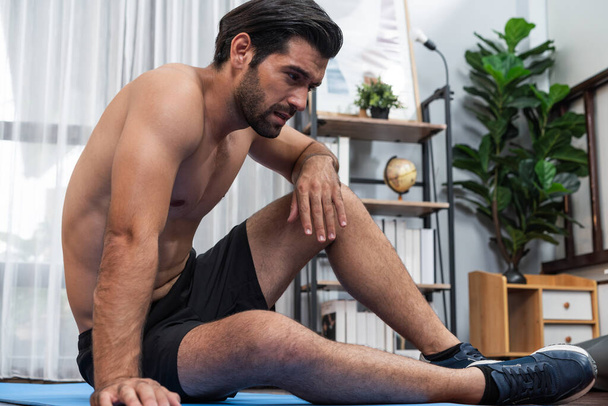 Athletischer und sportlicher Mann im Sitzen, auf Fitnessmatte liegend, nachdem er das Training zu Hause beendet hat, für einen fitten Körper und einen gesunden Lebensstil zu Hause. Fröhliches Heimtraining. - Foto, Bild