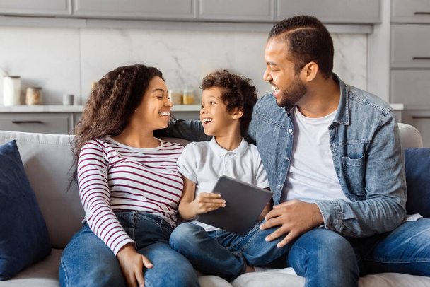 Famille heureuse afro-américaine composée d'une mère, d'un père et d'un jeune fils assis sur un canapé, partageant un moment de joie et de conversation, à l'aide d'une tablette numérique - Photo, image