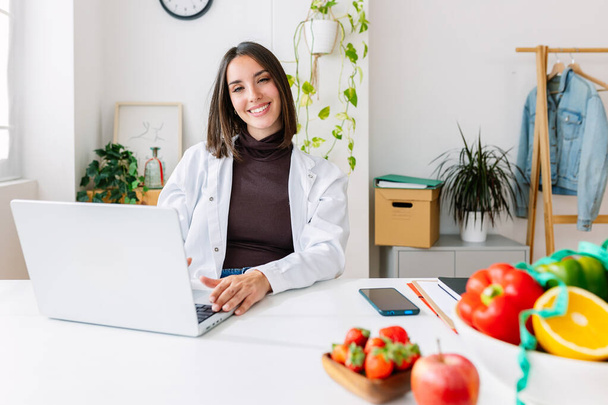 Smiling dietitian woman looking at camera sitting at hospital workplace. Nutrition, diet and professional healthcare worker concept - Photo, Image