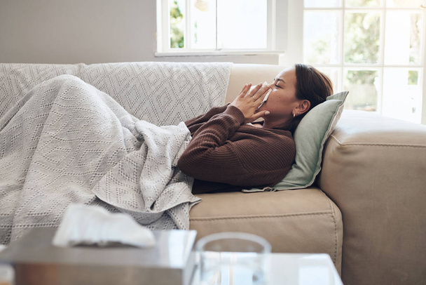 Chora kobieta, kichanie i dmuchanie nosa grypą, przeziębieniem lub chorobą na kanapie w salonie w domu. Młoda kobieta z tkanką bakterii, gorączki lub grypy w zmęczeniu lub odpoczynku na kanapie w salonie w domu. - Zdjęcie, obraz