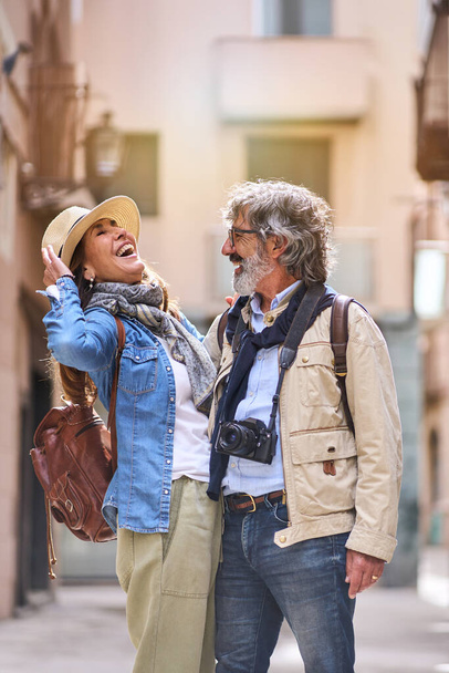 Κάθετα. Γελώντας λευκό ζευγάρι στέκεται αγκάλιασε στοργική στην τουριστική πόλη δρόμο. Οι ώριμοι αγαπούν τους ανθρώπους που απολαμβάνουν ρομαντικές διακοπές. Θετικές σχέσεις και αποδράσεις Σαββατοκύριακου άνοιξη - Φωτογραφία, εικόνα