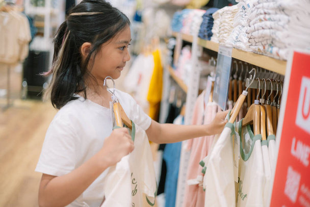 Egy fiatal lány ruhákat vásárol egy boltban. Egy inget tart a kezében, és nézi. - Fotó, kép