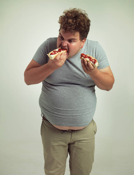 Plus size man, fast food and eating hotdogs for lunch in studio background for snack, hunger and craving. Male person, hungry and takeaway meal with ketchup or starving and enjoy while standing. - Photo, Image