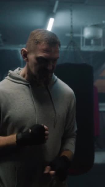 Sportowy bokser stoi w ciemnej sali bokserskiej i patrzy w kamerę. Mężczyzna wojownik w boksie bandaże rozciąga, uderza worek bokserski i przygotowuje się do treningu. Strzał pionowy - Materiał filmowy, wideo