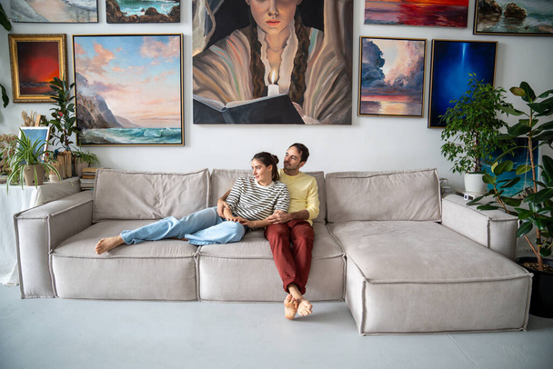 Uvolněný kreativní milující pár sedí na gauči a dívá se stranou, jak se něžně objímají v domácím uměleckém studiu. Šťastná žena s mužem objímající doma snění, plánování budoucnosti, vychutnat klid. - Fotografie, Obrázek