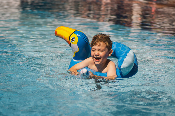 Chłopiec o radosnym wyrazie twarzy unosi się w basenie w słoneczny dzień, używając okręgu pływackiego zaprojektowanego tak, by wyglądał jak niebieski ptak.. - Zdjęcie, obraz