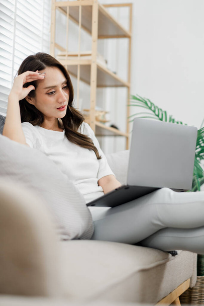 Mujer pensativa sentada en un sofá, trabajando en un ordenador portátil en un ambiente acogedor y bien iluminado. - Foto, imagen