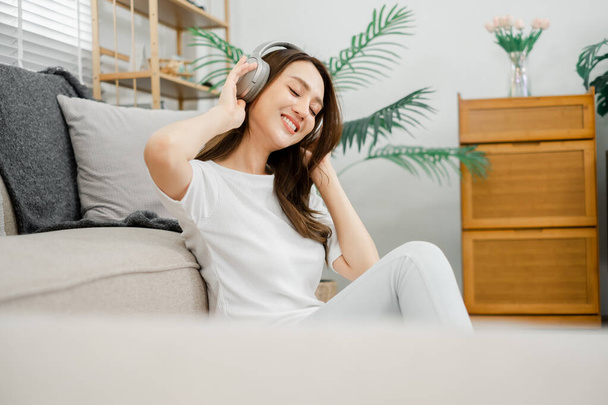 Radosna kobieta z zamkniętymi oczami czująca muzykę przez słuchawki, siedząca na kanapie w domu. - Zdjęcie, obraz