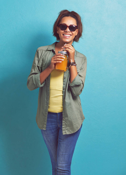 Mujer feliz, retrato y gafas de sol con zumo de naranja de moda para vitamina C o dieta en un fondo de estudio azul. Mujer, morena o modelo con sonrisa para bebida cítrica o bebida fresca. - Foto, Imagen