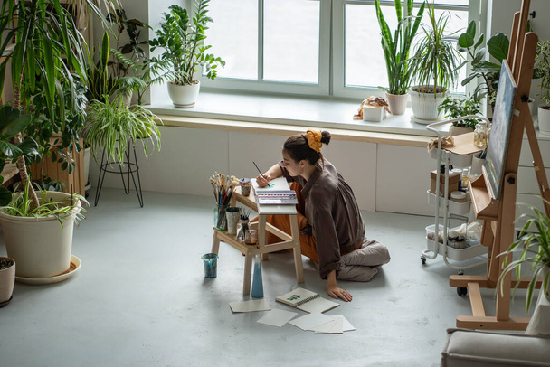 Creative woman artist sits on floor, painting in cozy art studio with houseplants. Drawing process with inspiration in craft workshop for stress relieve. Imagination, emotions on paper, brush strokes. - Photo, Image