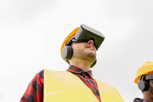 Niski kąt widzenia młodego dorosłego pracownika budowlanego w zestawie słuchawkowym VR, wyposażonego w sprzęt bezpieczeństwa. - Zdjęcie, obraz