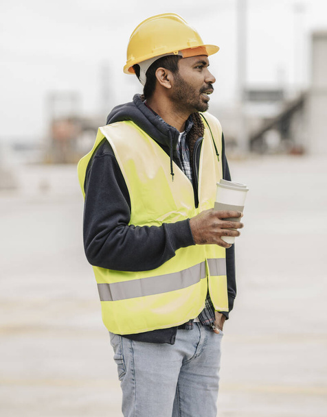 Młody pracownik budowlany z Azji Południowej trzymający filiżankę kawy, noszący kask bezpieczeństwa i kamizelkę na miejscu pracy. - Zdjęcie, obraz